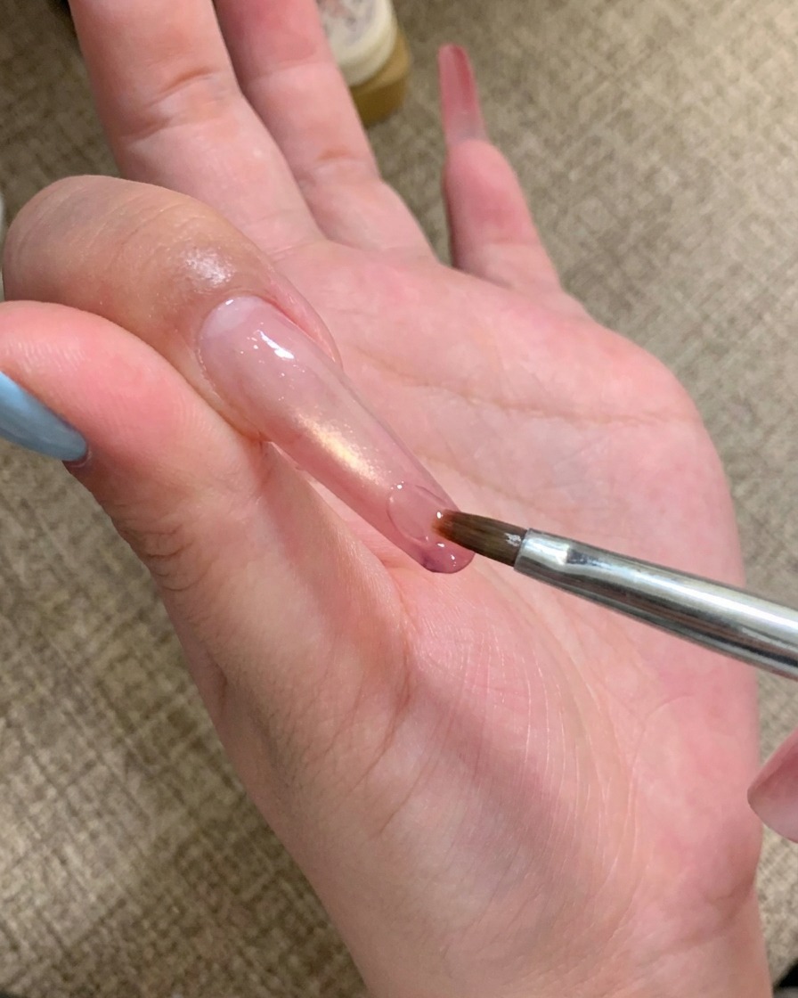 Un pennellino per nail art applica una goccia di gel adesivo trasparente sulla punta di ununghia lunga.