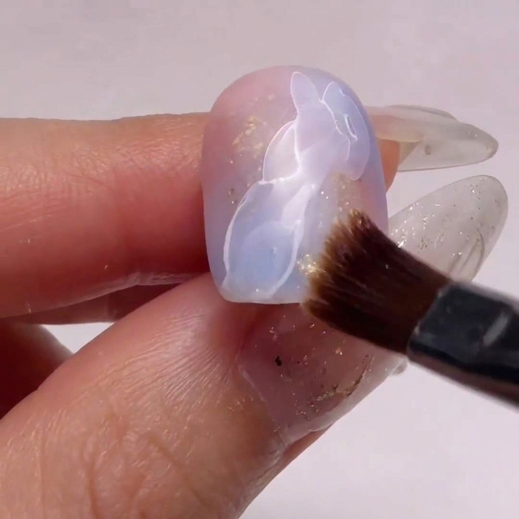 Primo piano di un piccolo pennello da unghie che usa alcol per sfumare un bordo di nail art gel bianca su base rosa e blu.