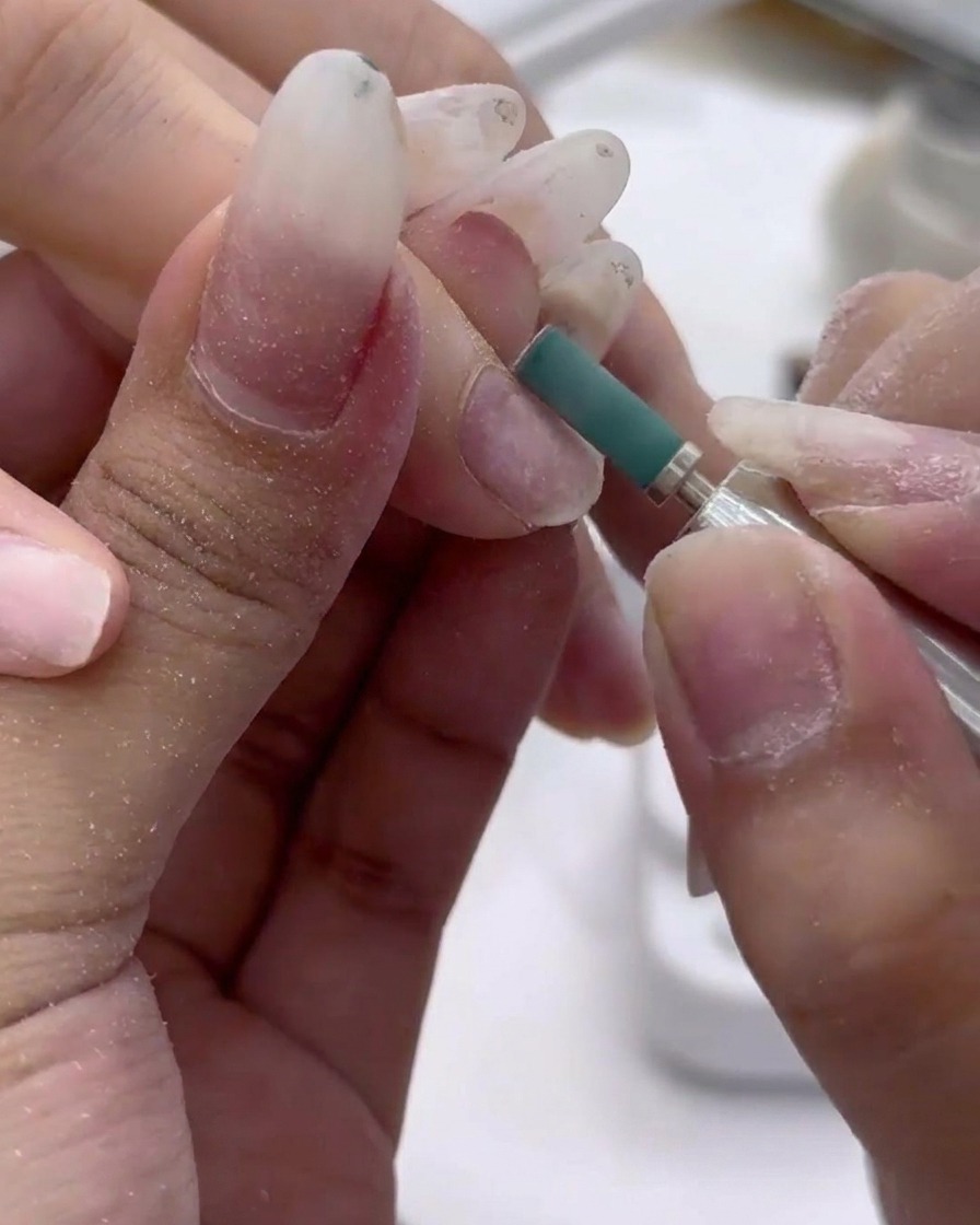 Primo piano di una punta abrasiva verde che leviga delicatamente la superficie di ununghia naturale.