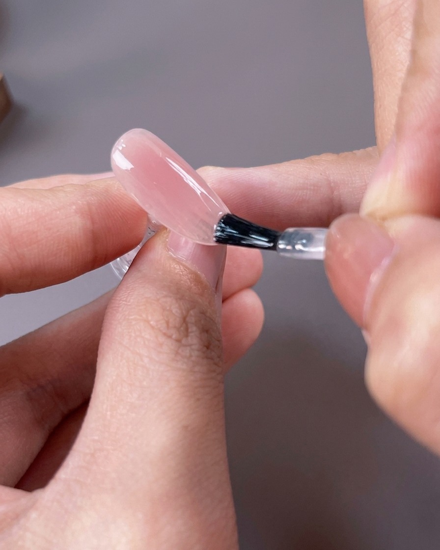Un pennello da unghie che applica uno smalto gel nude rosato traslucido sullintera superficie dellunghia.