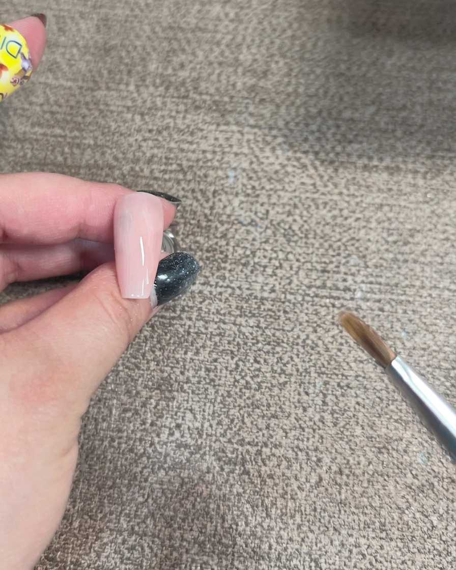 Primo piano di un pennello che applica gel iridescente con un motivo curvo su una base ungueale rosa chiaro.