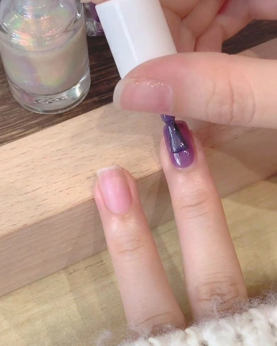 Un primo piano di un pennello che applica smalto gel viola profondo su ununghia naturale.