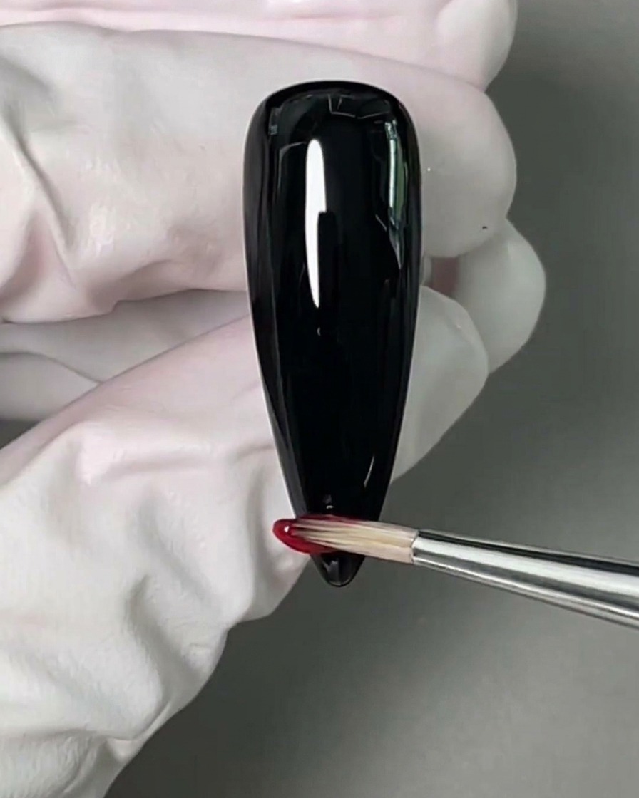 Un pennello da nail art applica uno smalto gel rosso vivace sulla punta di ununghia nera.
