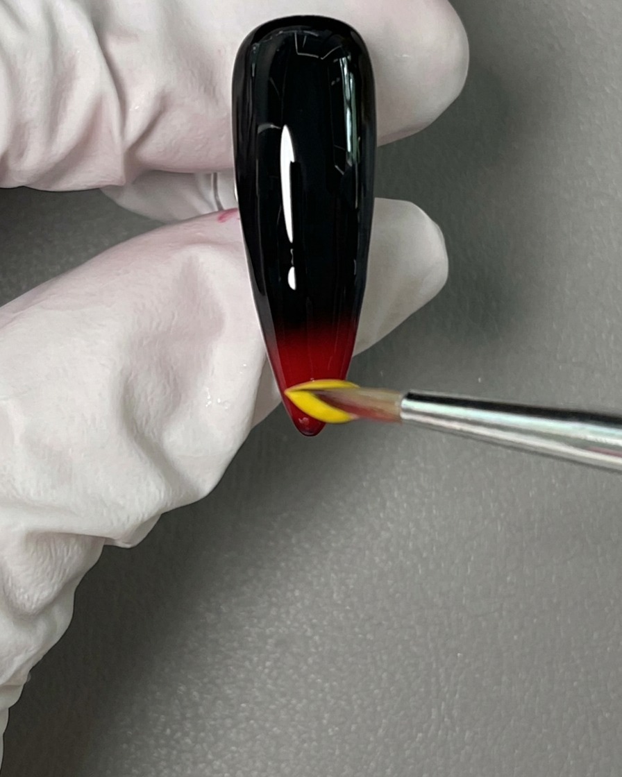 Un pennello di precisione posiziona una goccia di smalto gel giallo brillante sopra il rosso bagnato sulla punta dellunghia.