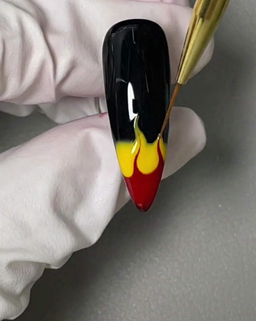 Un pennello liner a punta fine trascina verso lalto i gel bagnati rosso e giallo su ununghia nera per formare delle fiamme.