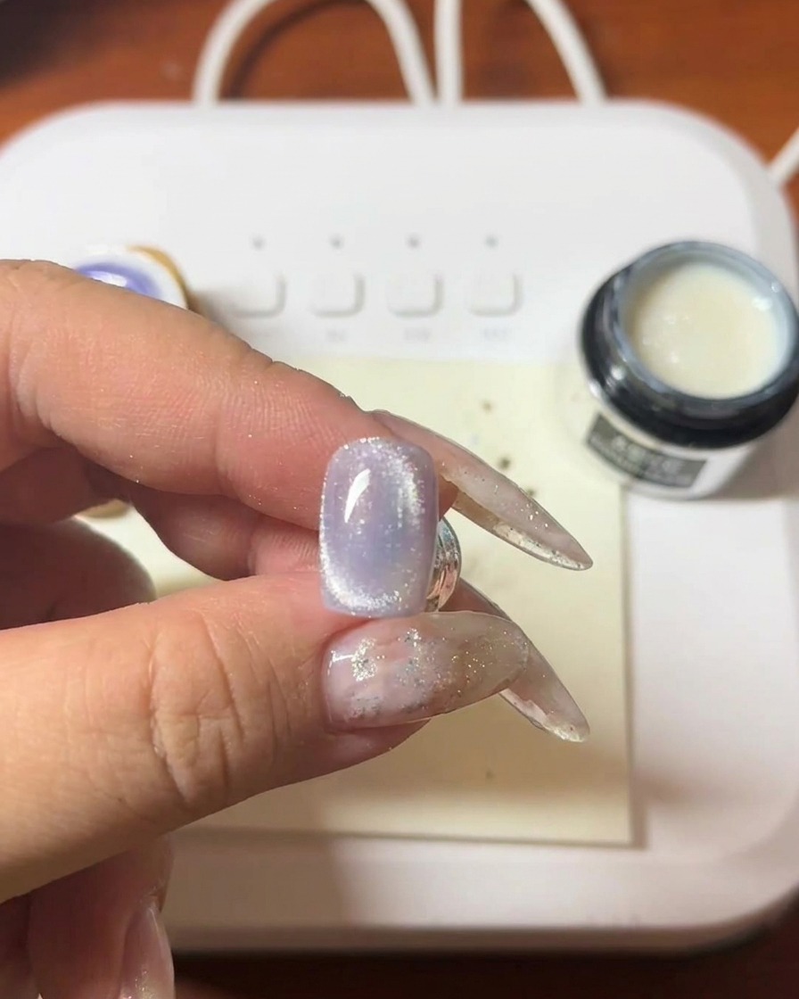 Primo piano di una mano che tiene una bacchetta magnetica sopra ununghia con gel cat-eye viola, mostrando la formazione della striscia luminosa.