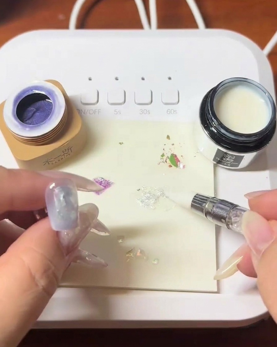 Un pennello che applica un blooming gel trasparente sopra lunghia decorata con conchiglie.