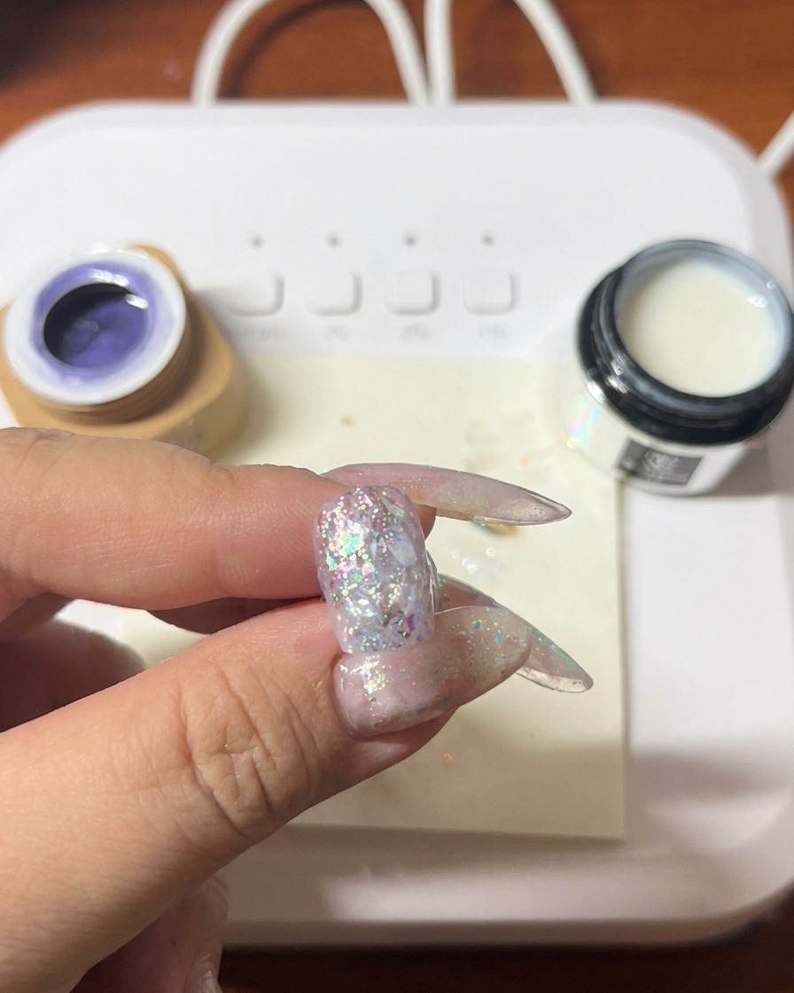 Un pennello che applica smalto gel viola su ununghia già decorata con glitter e conchiglie.
