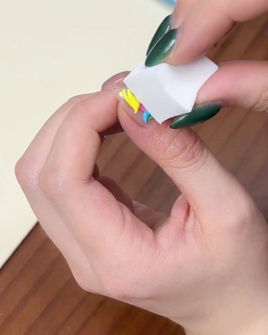 Primo piano di una mano che preme verticalmente una spugnetta bianca per trucco su ununghia con gocce di gel colorato.