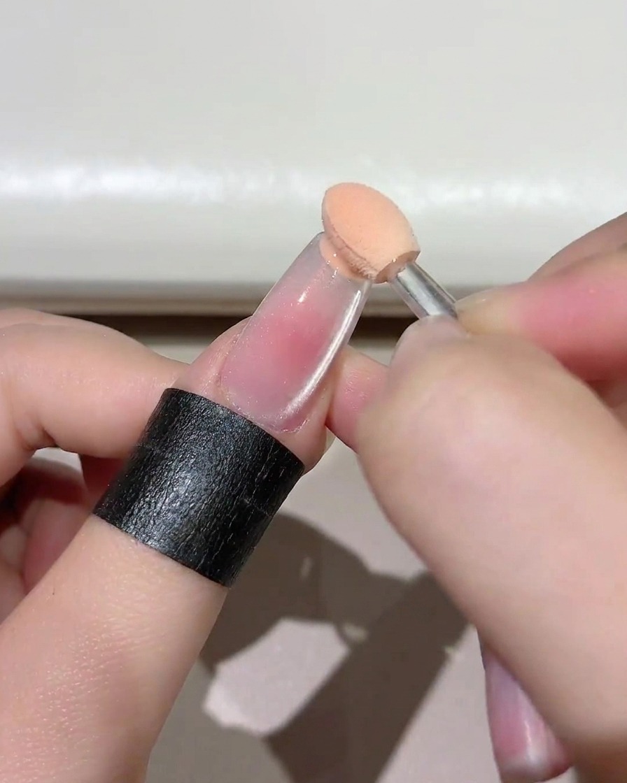 Primo piano di un piccolo applicatore a spugna che tampona gel rosato sul bordo libero di ununghia trasparente.