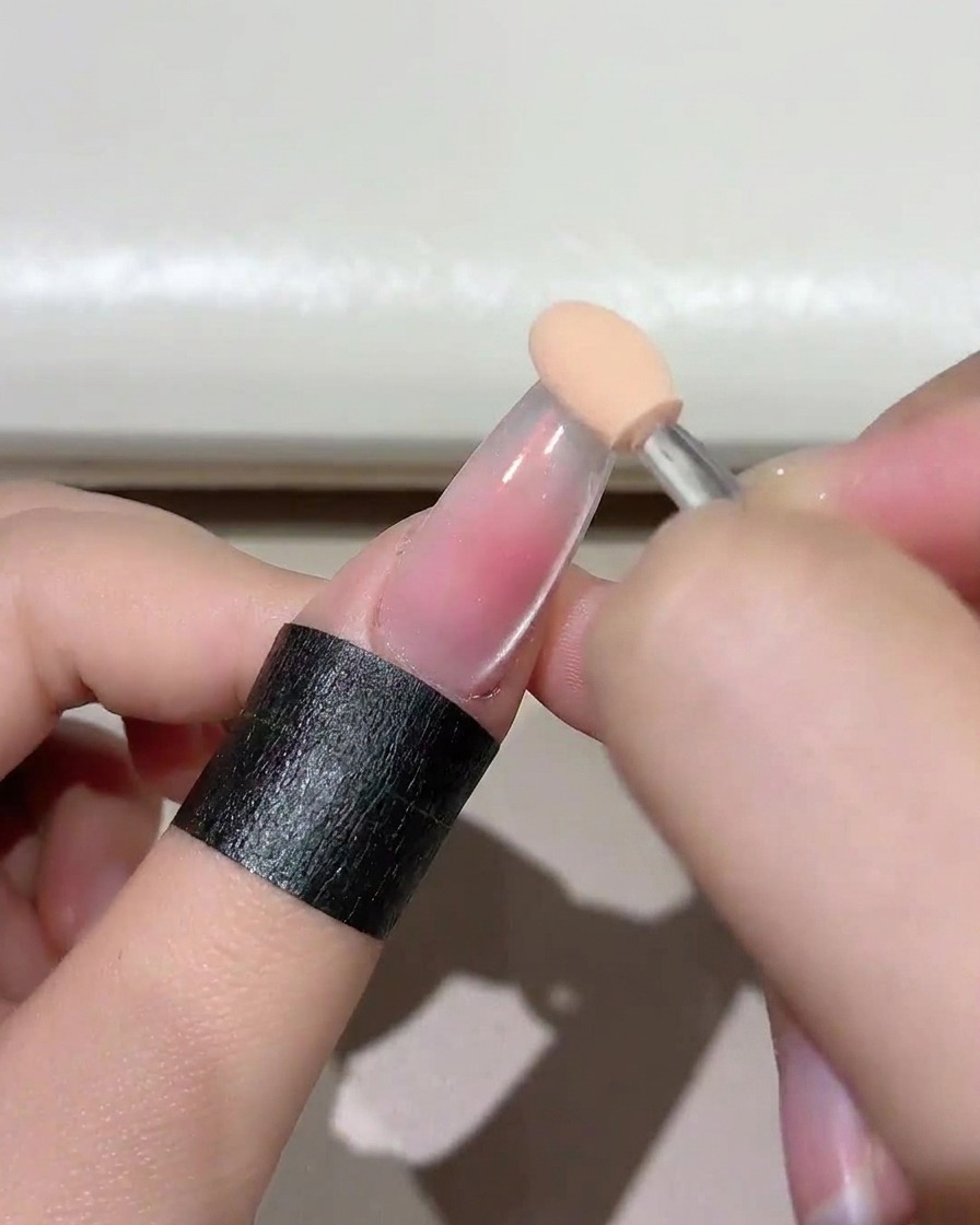 Primo piano di un applicatore a spugna che tampona un gel rosa blush delicato sulla punta dellunghia trasparente.