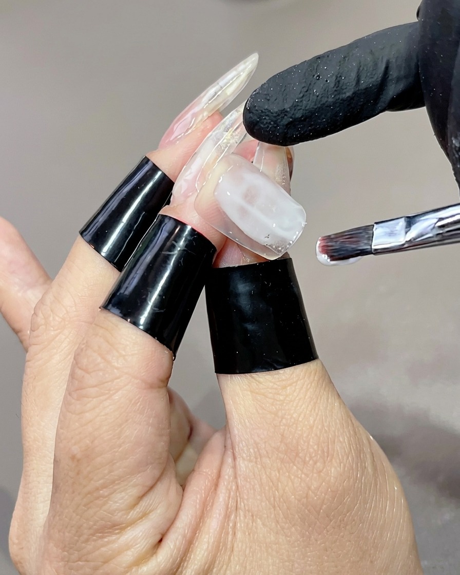 Una mano con tip lunghe e trasparenti, su cui un pennello piatto sta applicando una base gel bianco latte sullanulare.