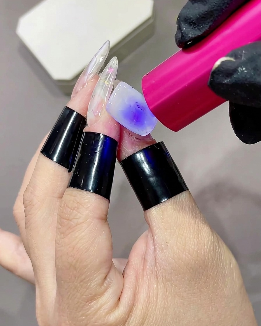 Primo piano di un dito con una base bianco latte che viene polimerizzata da una piccola torcia UV o LED rosa portatile.