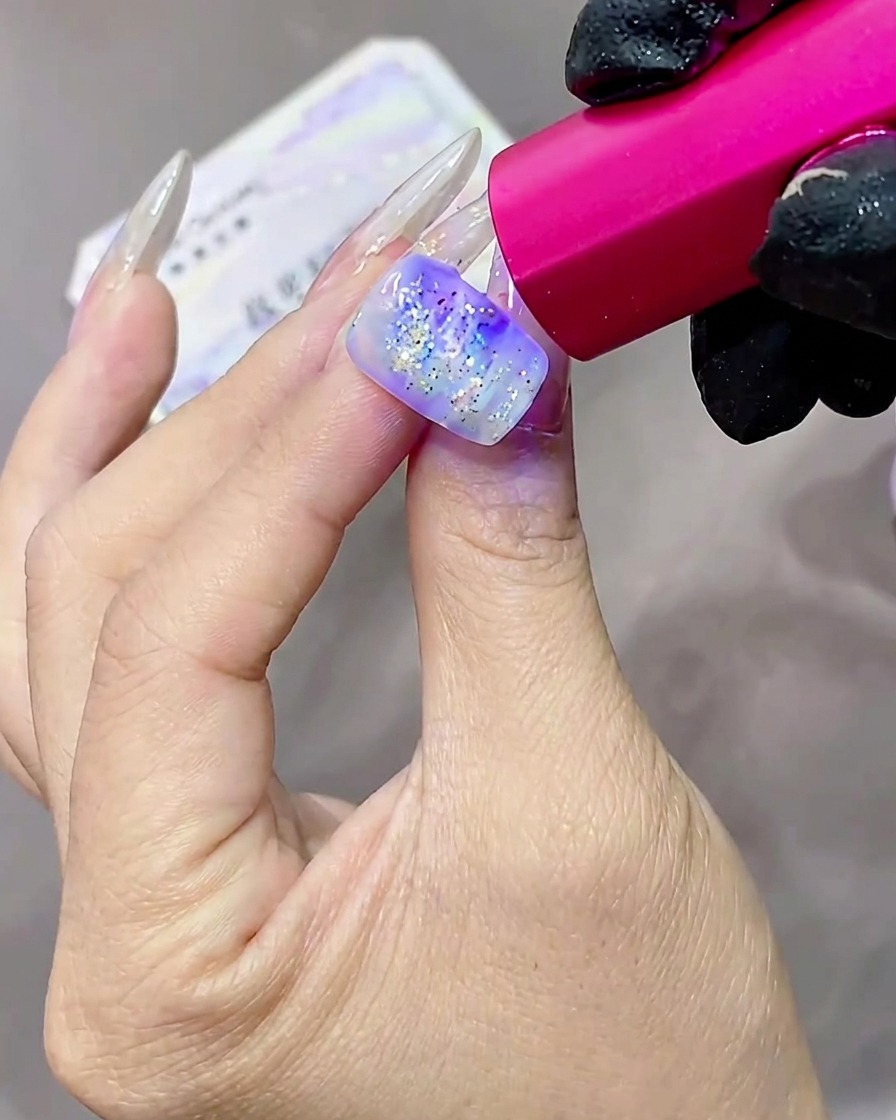 Una mano con unghie in gel glitterate e testurizzate che viene polimerizzata sotto una lampada UV dai toni rosa.
