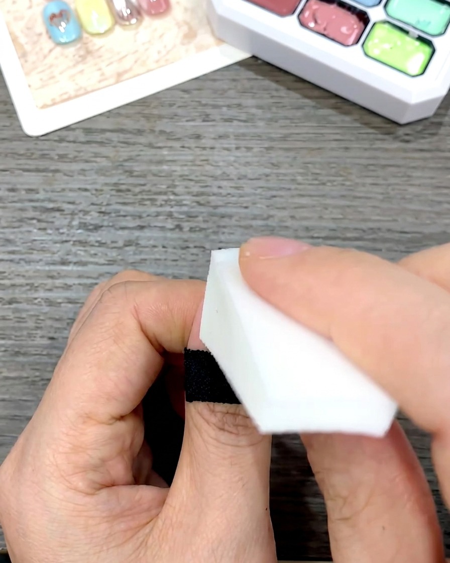 Una mano che usa una spugnetta cosmetica bianca a cuneo per picchiettare e sfumare delicatamente lo smalto gel giallo verso il basso su ununghia.