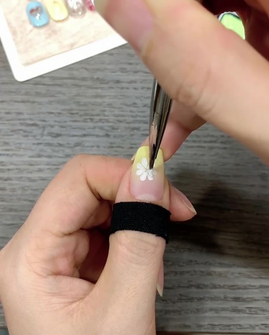 Vista ravvicinata di ununghia decorata con un pennello liner fine che applica un piccolo punto giallo brillante al centro di un design a margherita bianca.