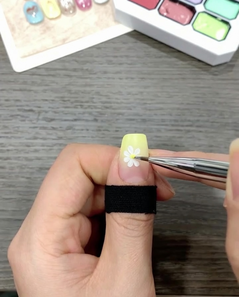 Un pennello che applica un top coat trasparente e lucido su ununghia decorata con un design a margherita su uno sfondo sfumato.