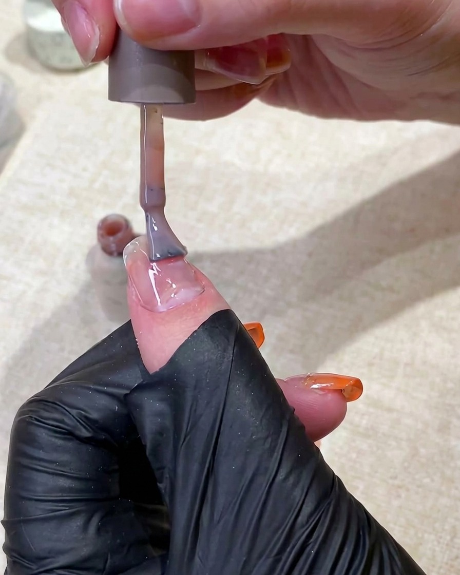 Una mano con guanto nero usa un pennello per stendere una base gel nude traslucida su ununghia.