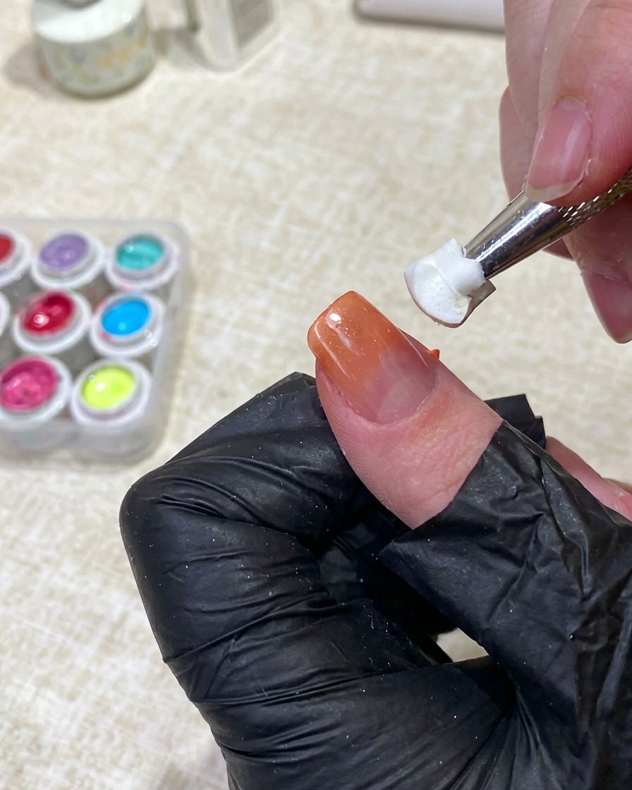 Uno strumento che tiene una spugnetta bianca pulita, tamponando leggermente il centro dellunghia per diffondere il gel arancione sulla base nude.