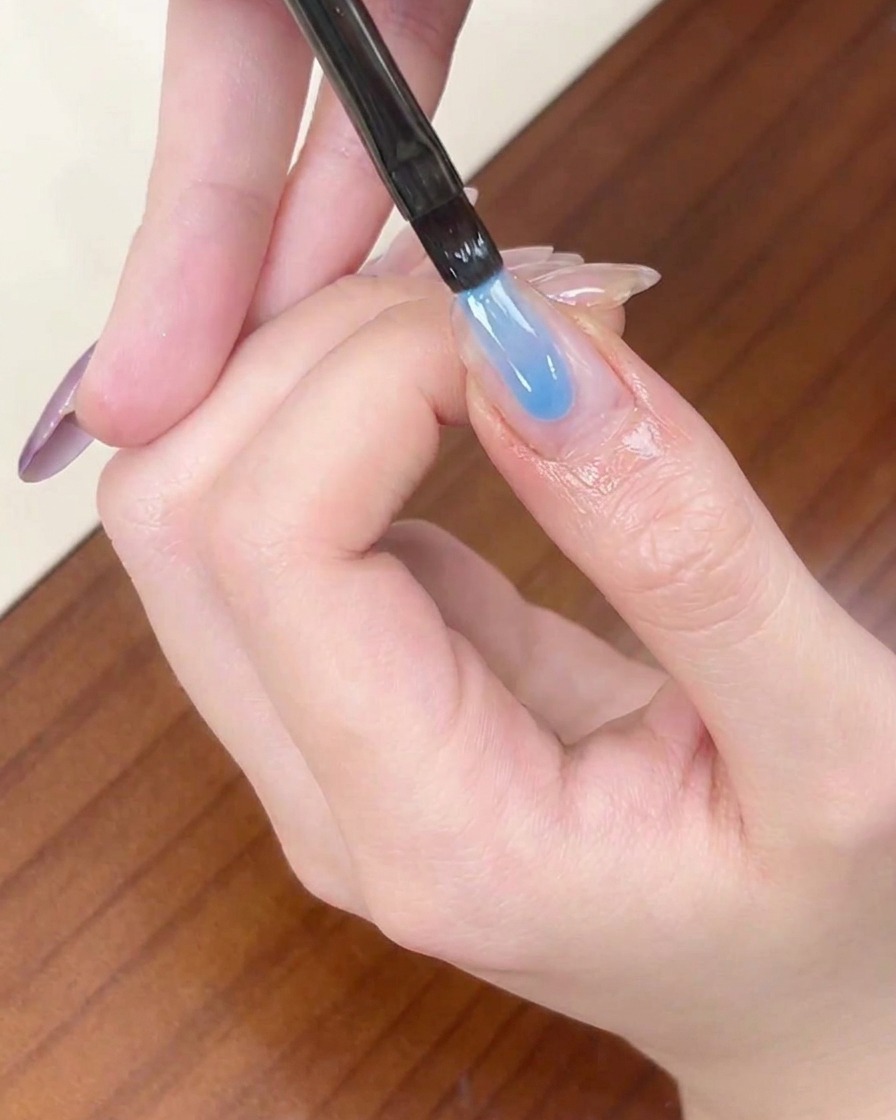 Un pennello liner applica una striscia di smalto gel azzurro semitrasparente sulla punta di ununghia a mandorla.