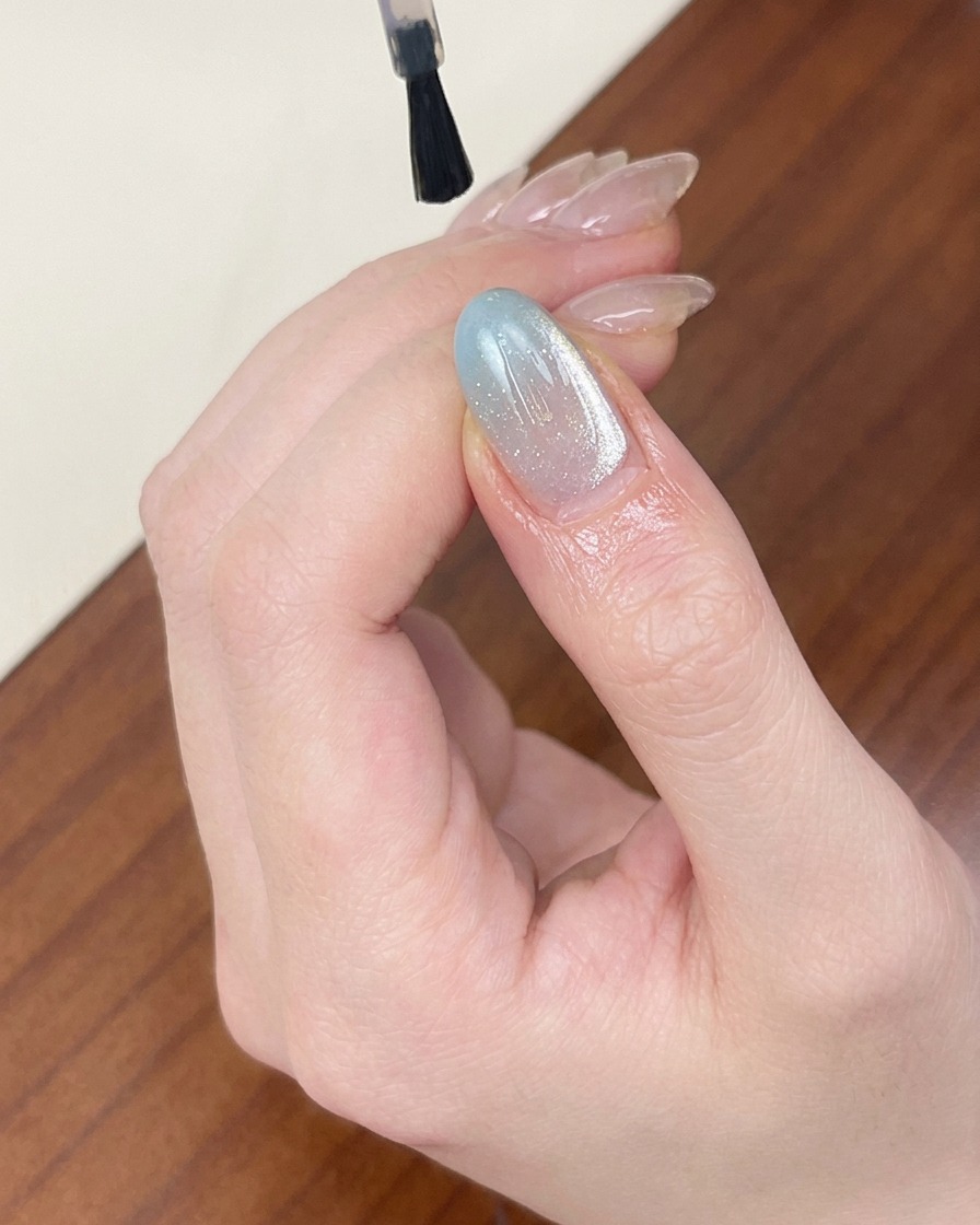 Un pennello applica un top coat trasparente e lucido su ununghia in gel cat-eye azzurra e scintillante.