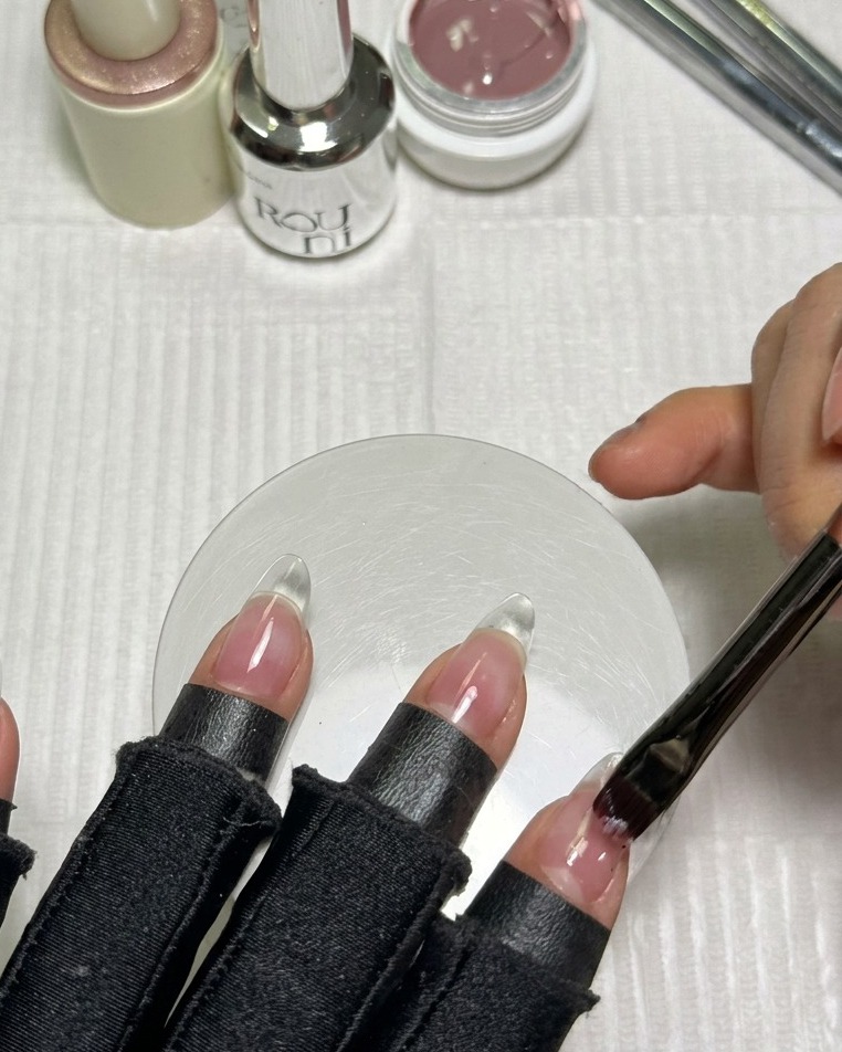 Un pennello applica uno strato uniforme di gel nude rosato su una tip a mandorla trasparente.