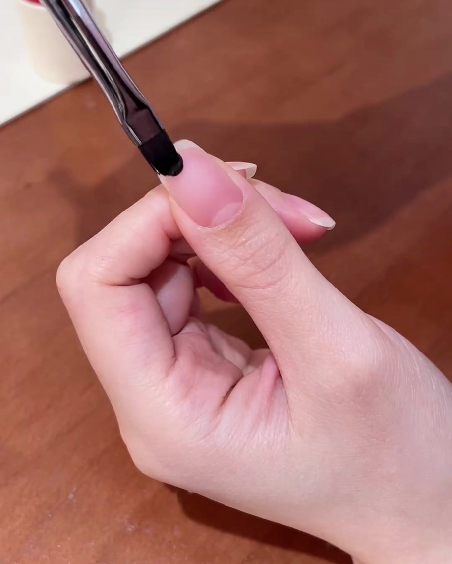 Primo piano di un pennello che applica una base gel trasparente su ununghia naturale pulita.