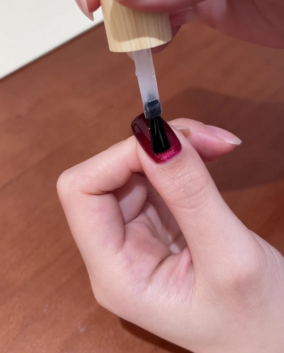 Una vista ravvicinata di una mano con top coat appena applicato su unghie cat-eye rosso scuro, inserita in una lampada UV per il passaggio finale.