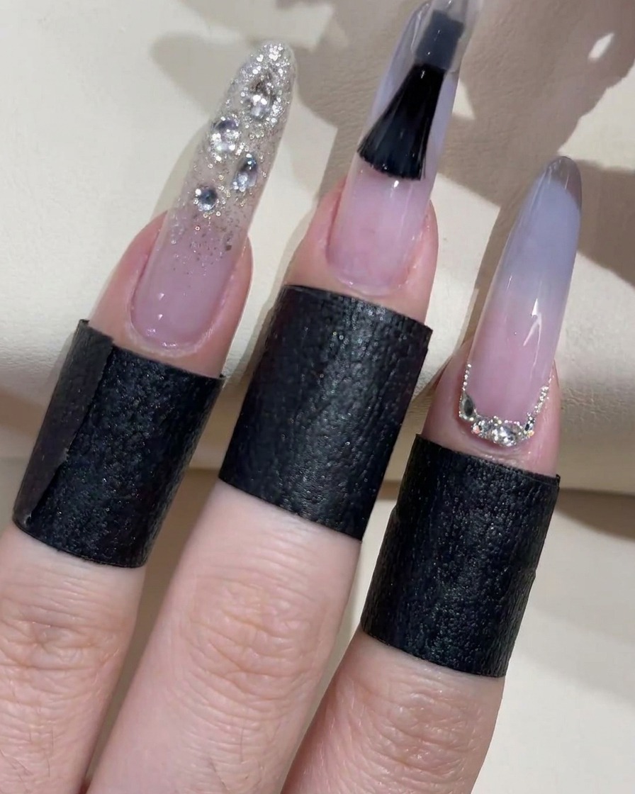 Un primo piano di un pennello per smalto nero che applica attivamente un top coat trasparente su ununghia blu ombré, con le dita adiacenti che mostrano glitter argento e decorazioni con strass.