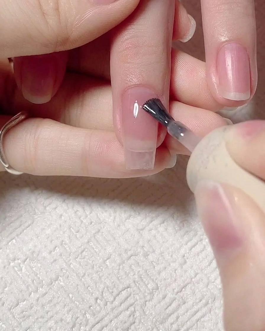 Primo piano di un pennello che applica un gel jelly rosa trasparente su ununghia naturale pulita e preparata.