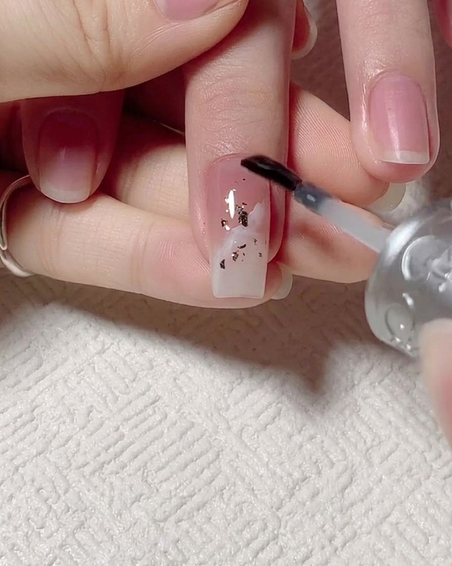 Un pennello che applica base gel trasparente sopra ununghia con design a foglia oro e onde.