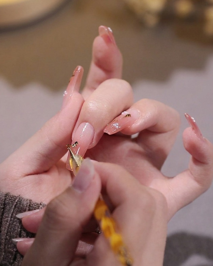 Una mano che usa un dotting tool per posizionare minuscole scaglie di foglia oro su ununghia dipinta di rosa.