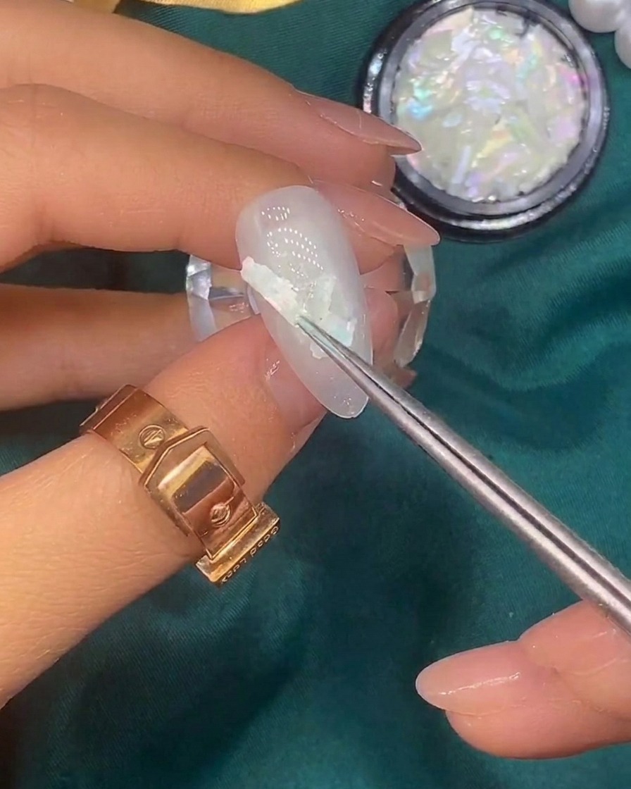 Un pennello applica minuscoli fiocchi di foglia doro sulla superficie di ununghia già decorata con conchiglie.