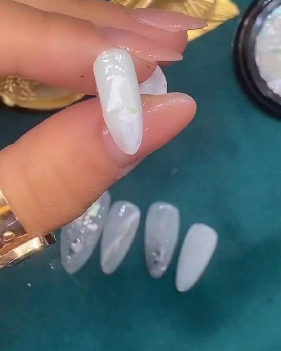 Pinzette che posizionano frammenti decorativi di conchiglia su ununghia coperta di gel bianco traslucido umido.