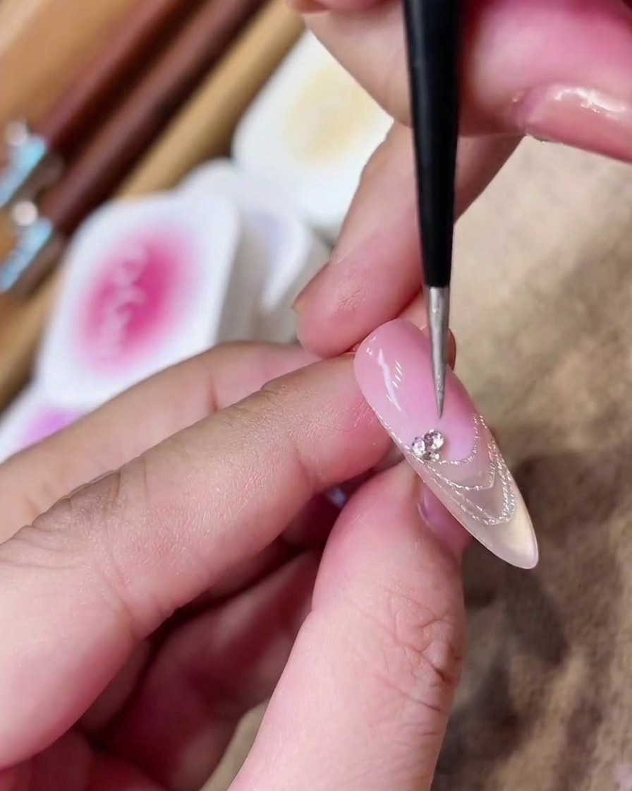 Un pennello da dettaglio posiziona un cluster di piccoli strass scintillanti sulla linea del sorriso di una French manicure.