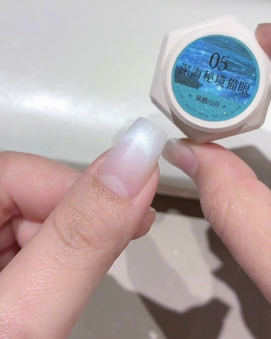 Mano che tiene un barattolino di gel cat-eye blu ottanio accanto a ununghia preparata con punta bianca.