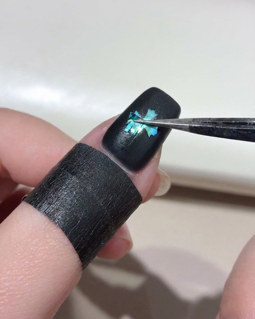 Pinzette che rimuovono lo stencil a croce dallunghia nera, rivelando il design cat-eye blu sottostante.