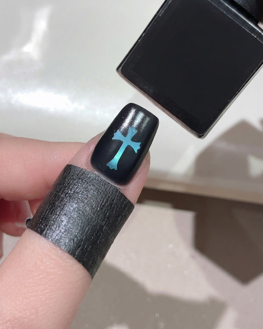 Pennello che applica un top coat lucido su ununghia nera con design a croce blu.