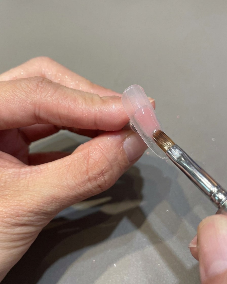 Un pennello professionale applica uno smalto gel rosa trasparente su una superficie ungueale preparata.