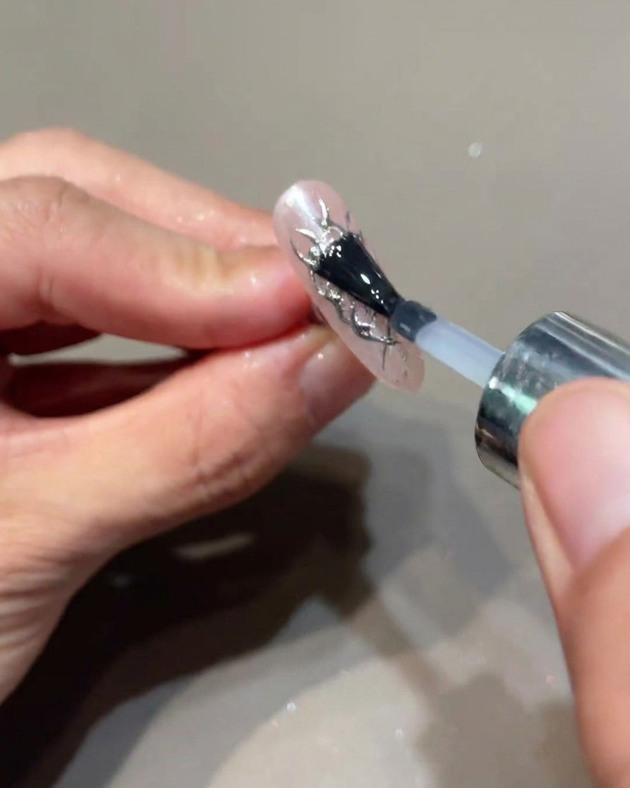Un pennello applica uno strato finale di top coat trasparente su ununghia decorata con fiamme argento.