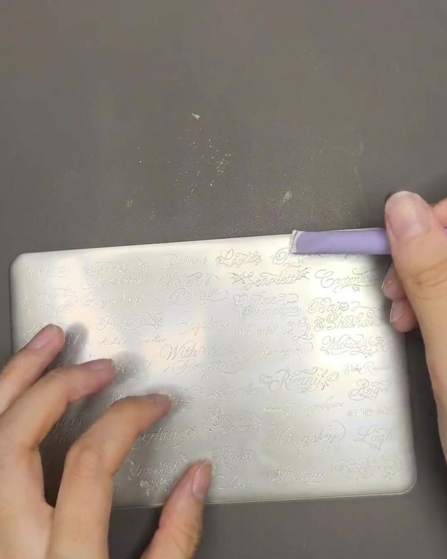 Un primo piano che mostra smalto per stamping bianco steso su una piastra in metallo con un raschietto viola.