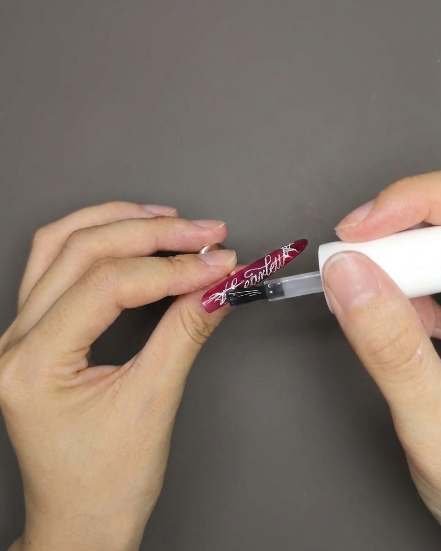 Un pennello che applica un top coat trasparente e lucido su ununghia rosso cat eye con scritte inglesi bianche stampate.