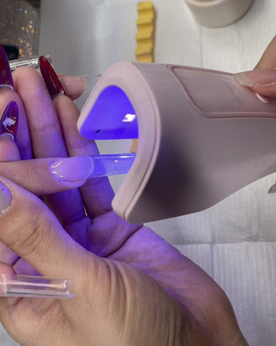 Una nail artist che tiene una piccola lampada LED rosa portatile sopra unestensione quadrata in gel trasparente per polimerizzarla.