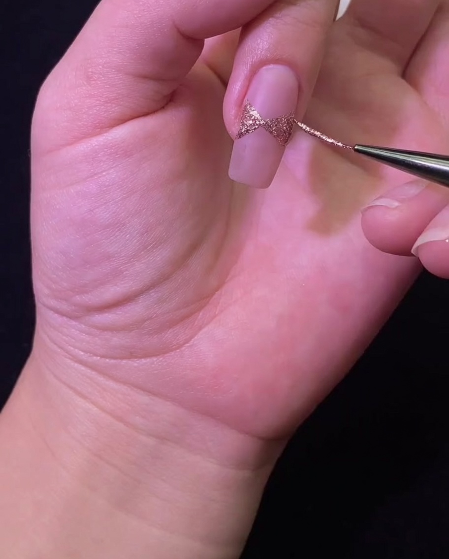 Vista ravvicinata di un pennello liner che perfeziona i bordi di un fiocco in glitter oro rosa dipinto su una base nude.