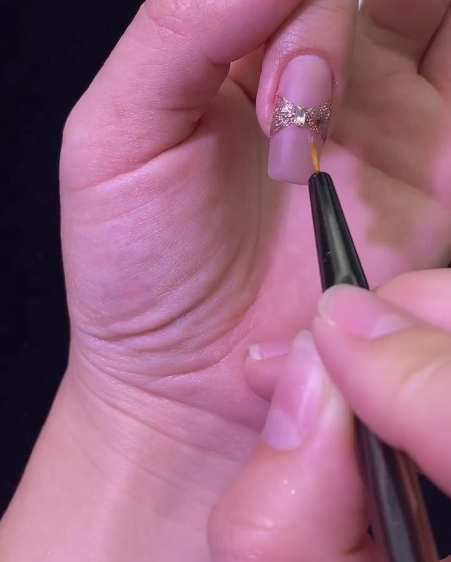 Un pennello liner che applica un top coat lucido per sigillare il fiocco in glitter oro rosa e lo strass centrale.