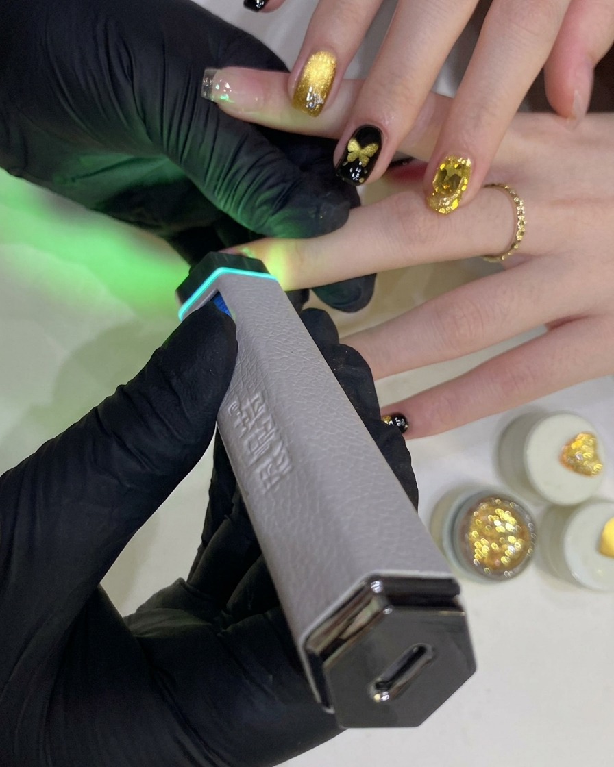 Una mano guantata tiene una lampada UV/LED portatile sopra ununghia con gel nero.
