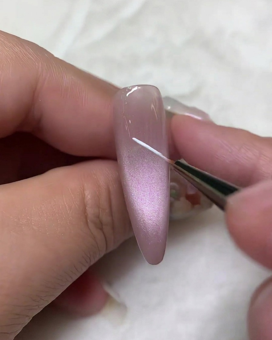 Vista ravvicinata di un pennello liner fine che applica una sottile linea bianca verticale al centro di ununghia in gel cat-eye color malva.