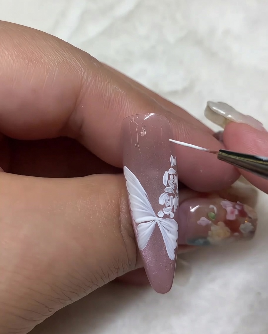 Primo piano che mostra un pennello liner che aggiunge piccoli punti luce bianchi attorno a un design con farfalla e rosa su ununghia nude.