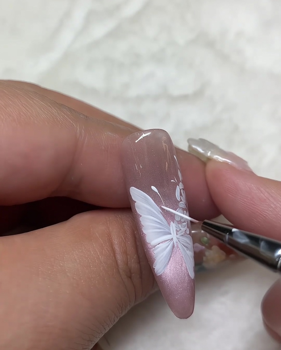 Primo piano di ununghia con un design a rosa bianca, mentre un pennello liner viene usato per eseguire gli ultimi ritocchi sui dettagli dei petali.