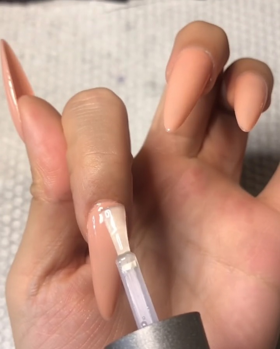 Primo piano di un pennello che applica con cura un top coat trasparente temperato vicino alla zona della cuticola di ununghia a mandorla nude.